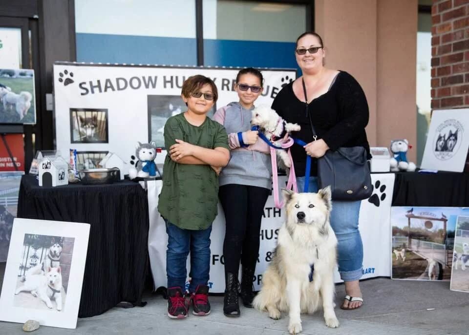 A woman and two children wearing sunglasses pose with a white Husky at an adoption event
