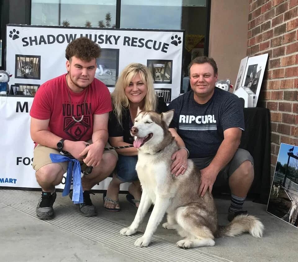 A family of three poses with a happy brown-and-white Husky in front of the Shadow Husky Rescue banner