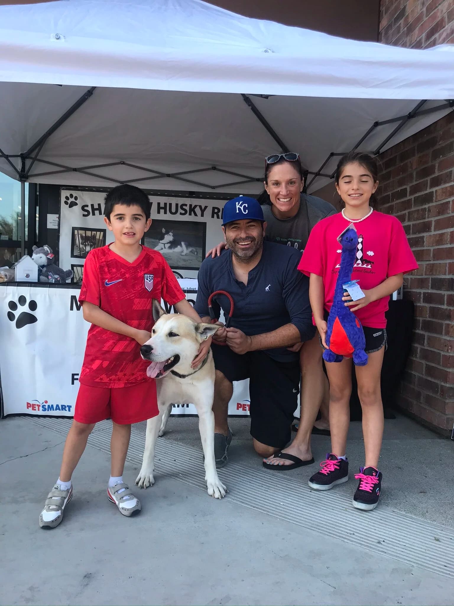 A happy family of four poses with a white husky mix at an outdoor adoption event