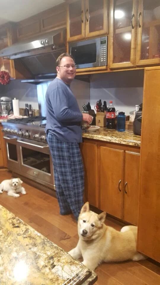 A man cooking in a kitchen while a golden Husky and a small white dog rest on the floor nearby
