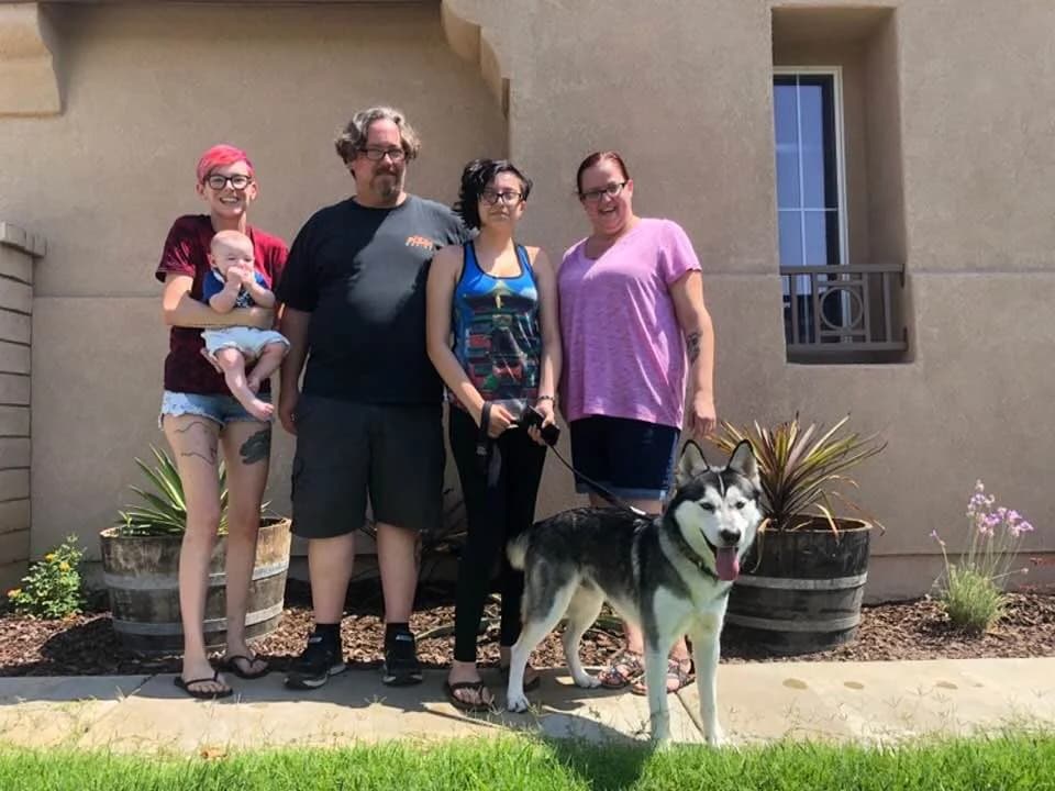 A group of four adults poses outside a home with a black-and-white Husky on a leash