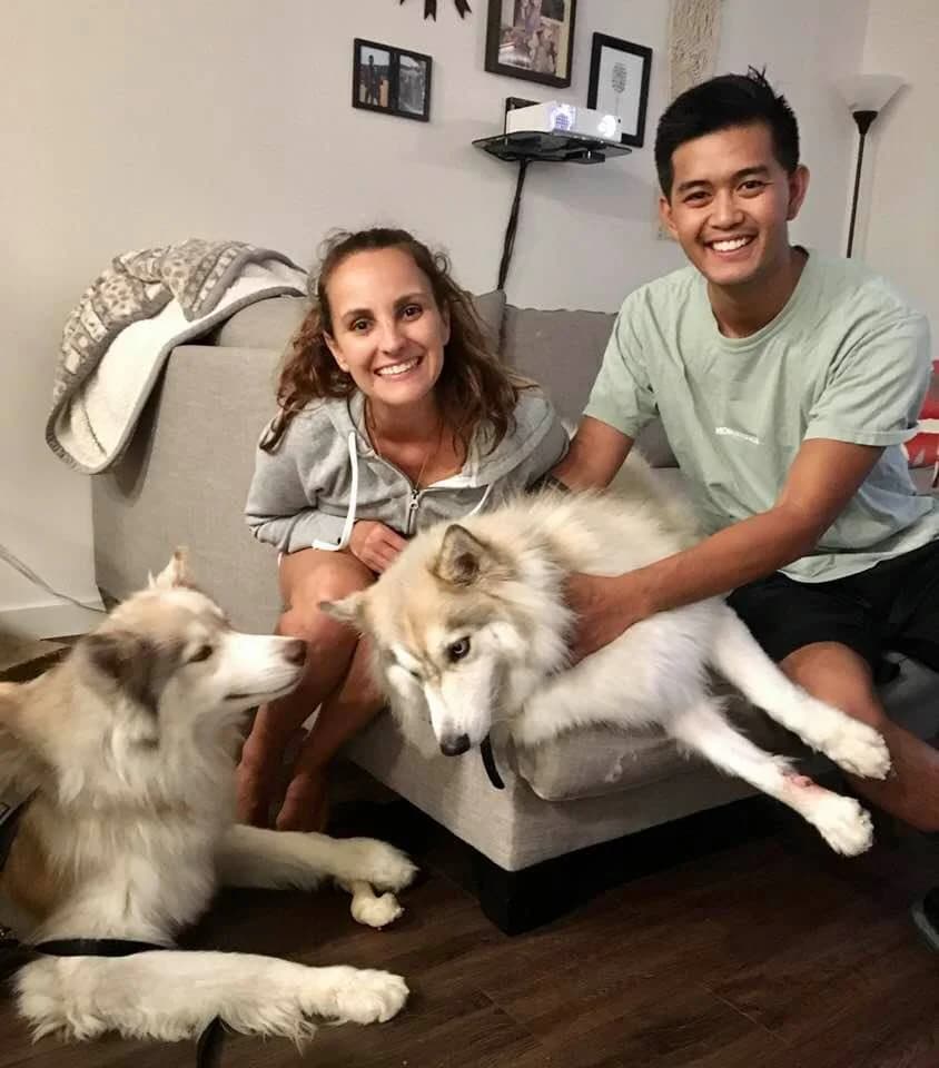 A smiling woman and young man sit on a couch surrounded by two fluffy Huskies