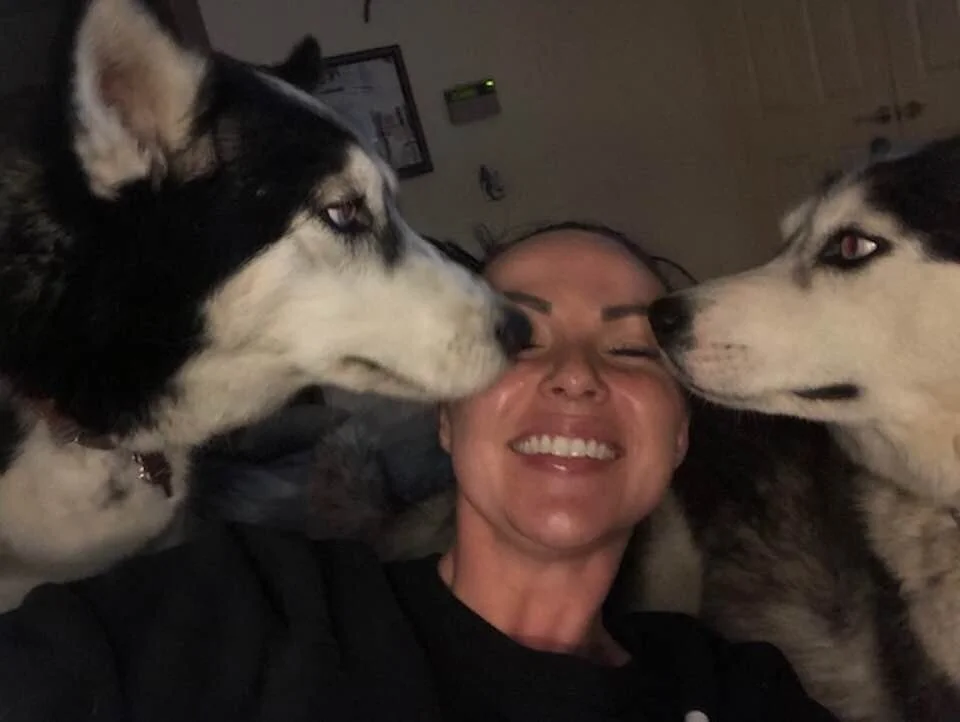 A woman laughing as two Siberian Huskies simultaneously lick both sides of her face