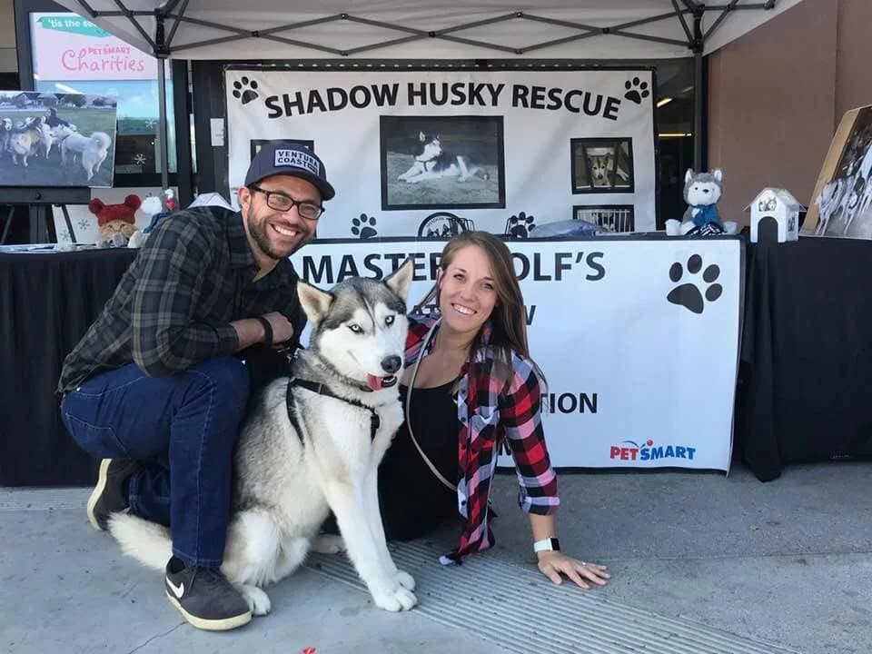 A smiling couple kneels with a blue-eyed Siberian Husky at a Shadow Husky Rescue adoption event booth