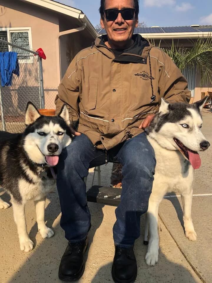 A man in sunglasses sits outdoors flanked by two Huskies in a residential setting