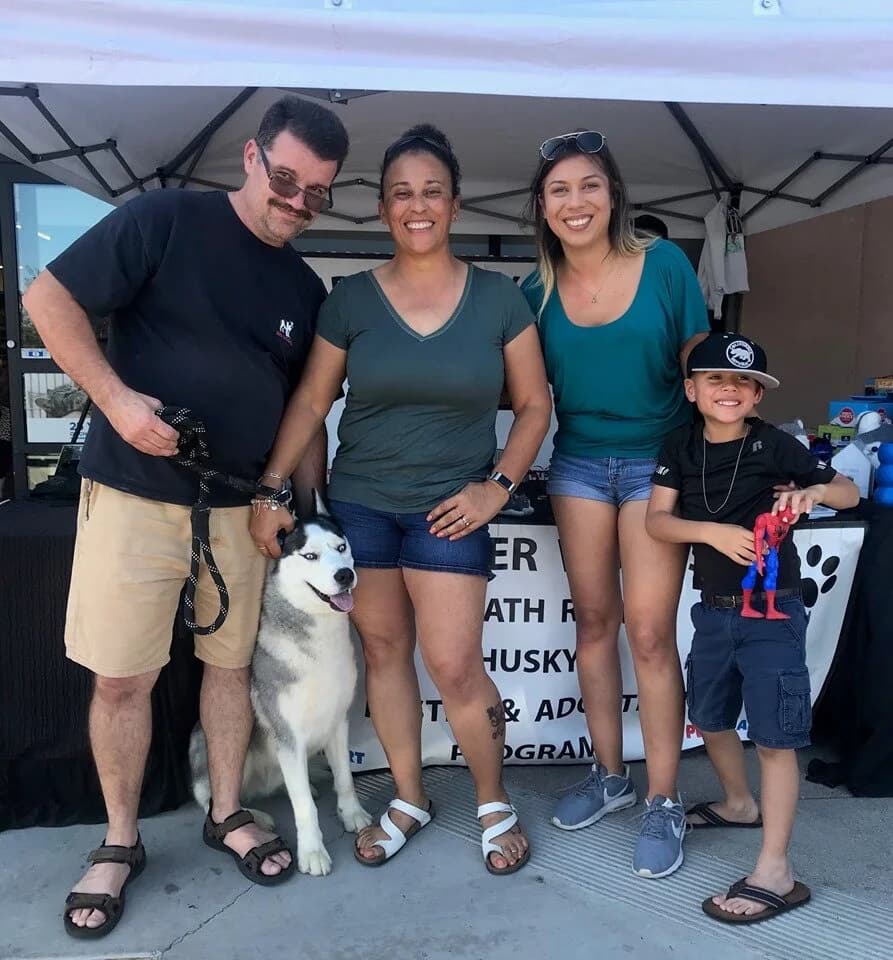 A smiling group of three adults and a child pose with a black-and-white husky at a rescue adoption event booth