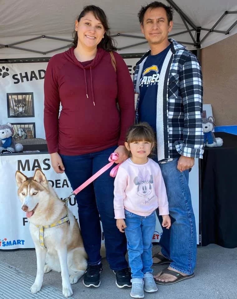 A family of three stands with a red husky on a leash at an outdoor adoption event
