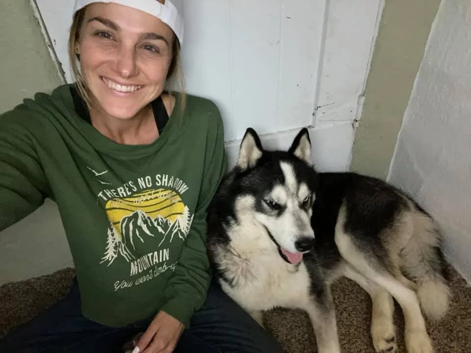 A smiling woman in a green sweatshirt takes a selfie with a black-and-white husky dog