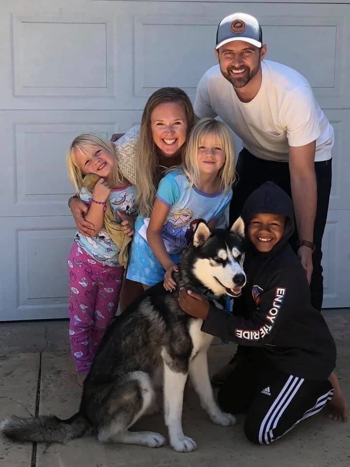 A happy family of five posing together with a black and white Husky in front of a garage door