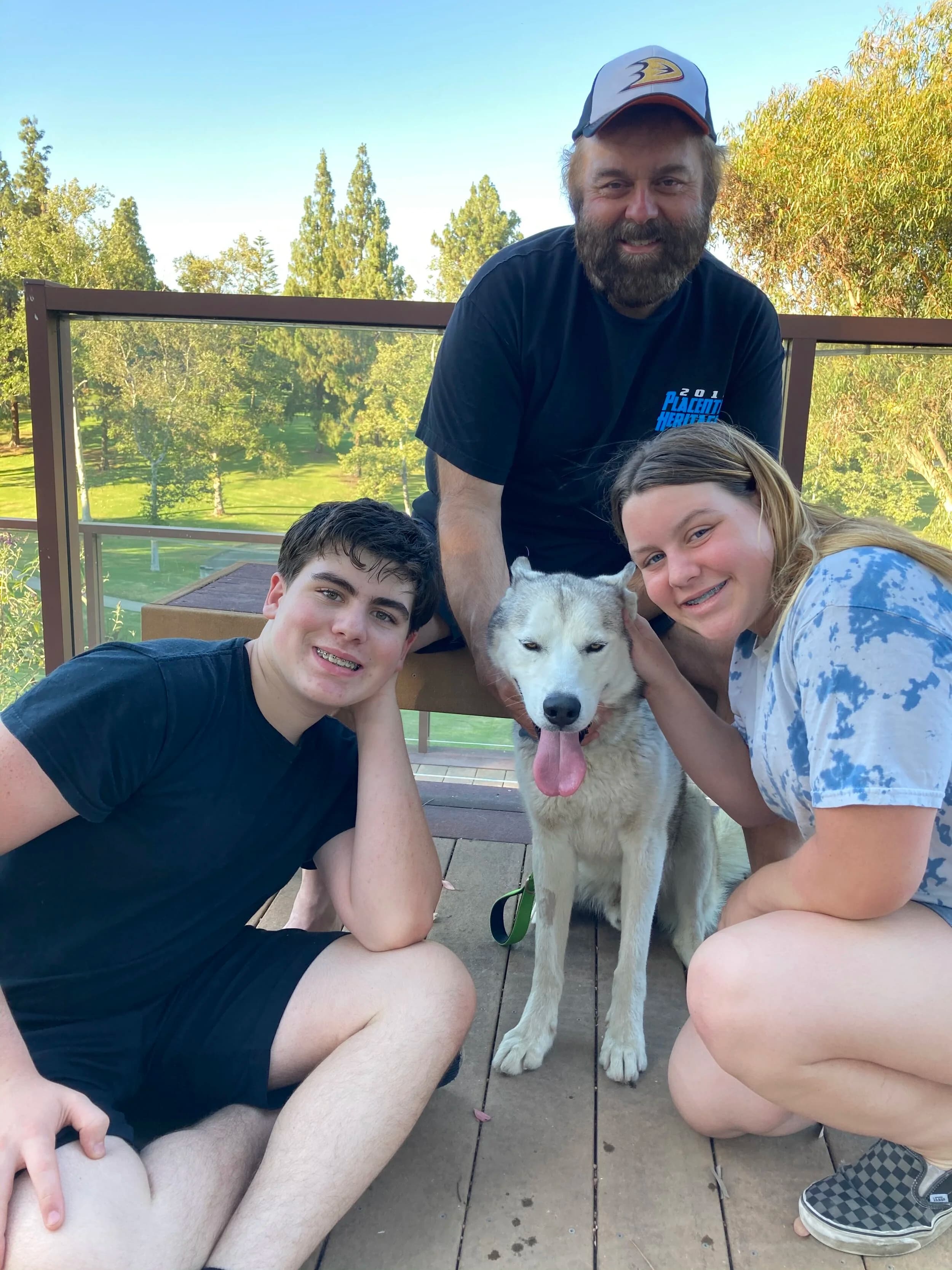 A smiling family of three posing outdoors on a deck with a happy white and grey Husky