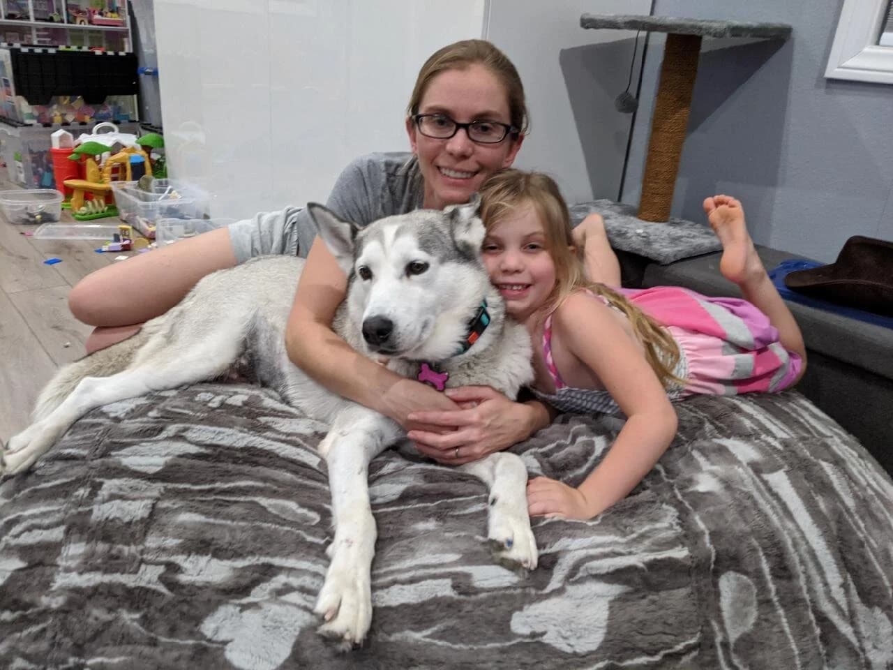 A mother and young daughter lying on a dog bed hugging and smiling with a grey and white Husky