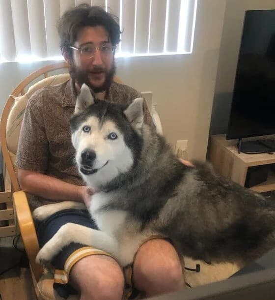 A young man sitting in a rattan chair with a large grey and white Husky lounging across his lap