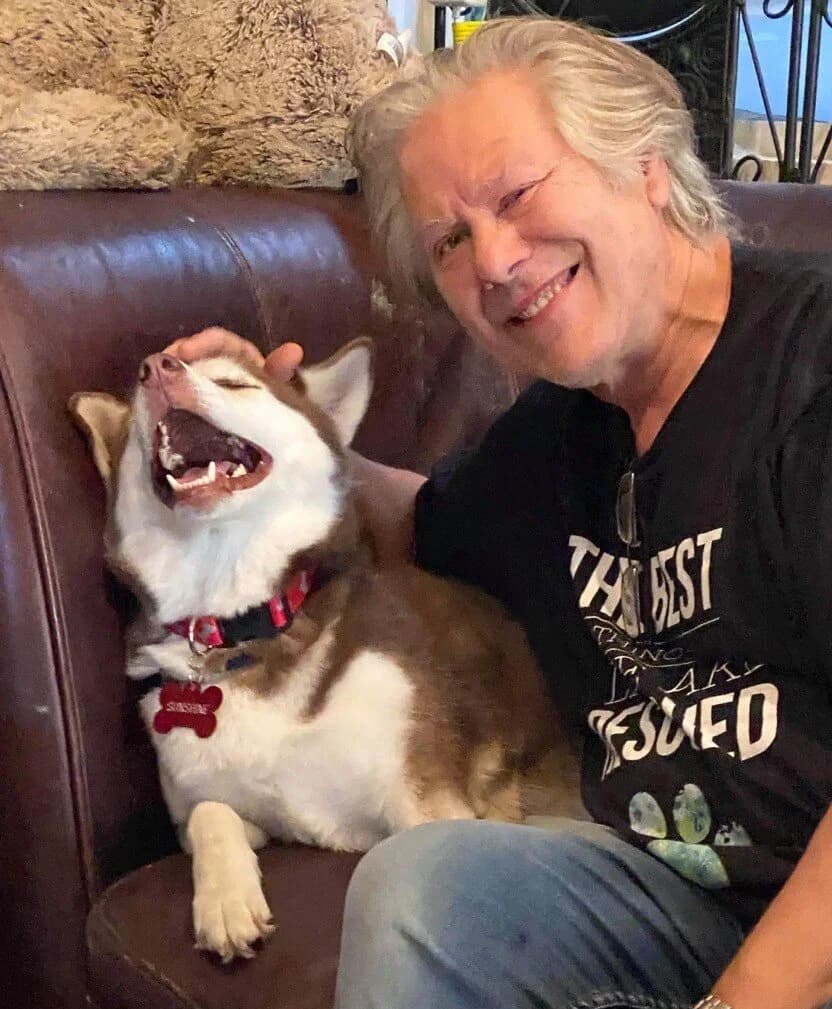 An older man laughing joyfully while a happy red and white Husky yawns widely beside him