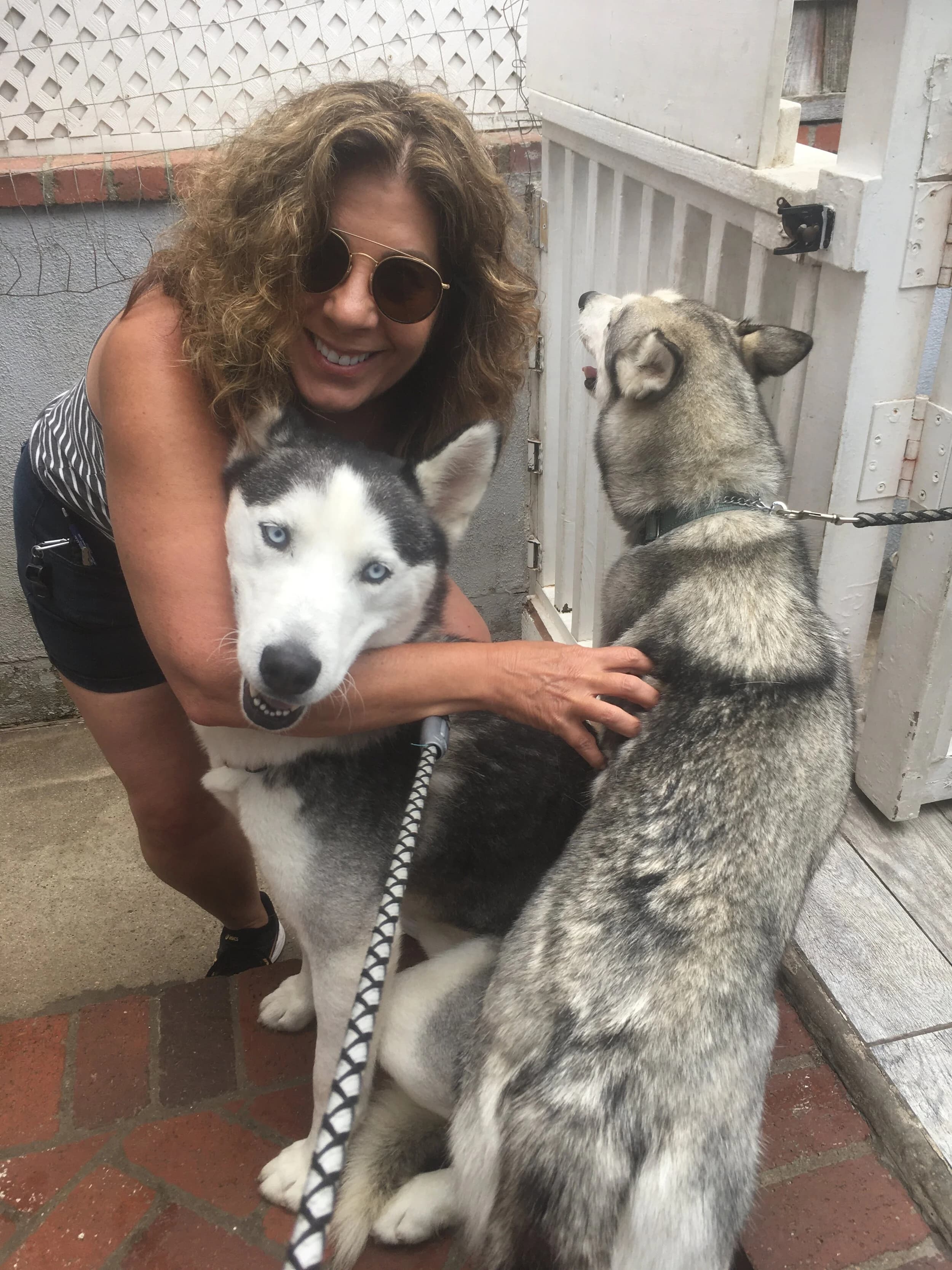 A smiling woman with sunglasses crouching to pet two Siberian Huskies on a tiled outdoor patio