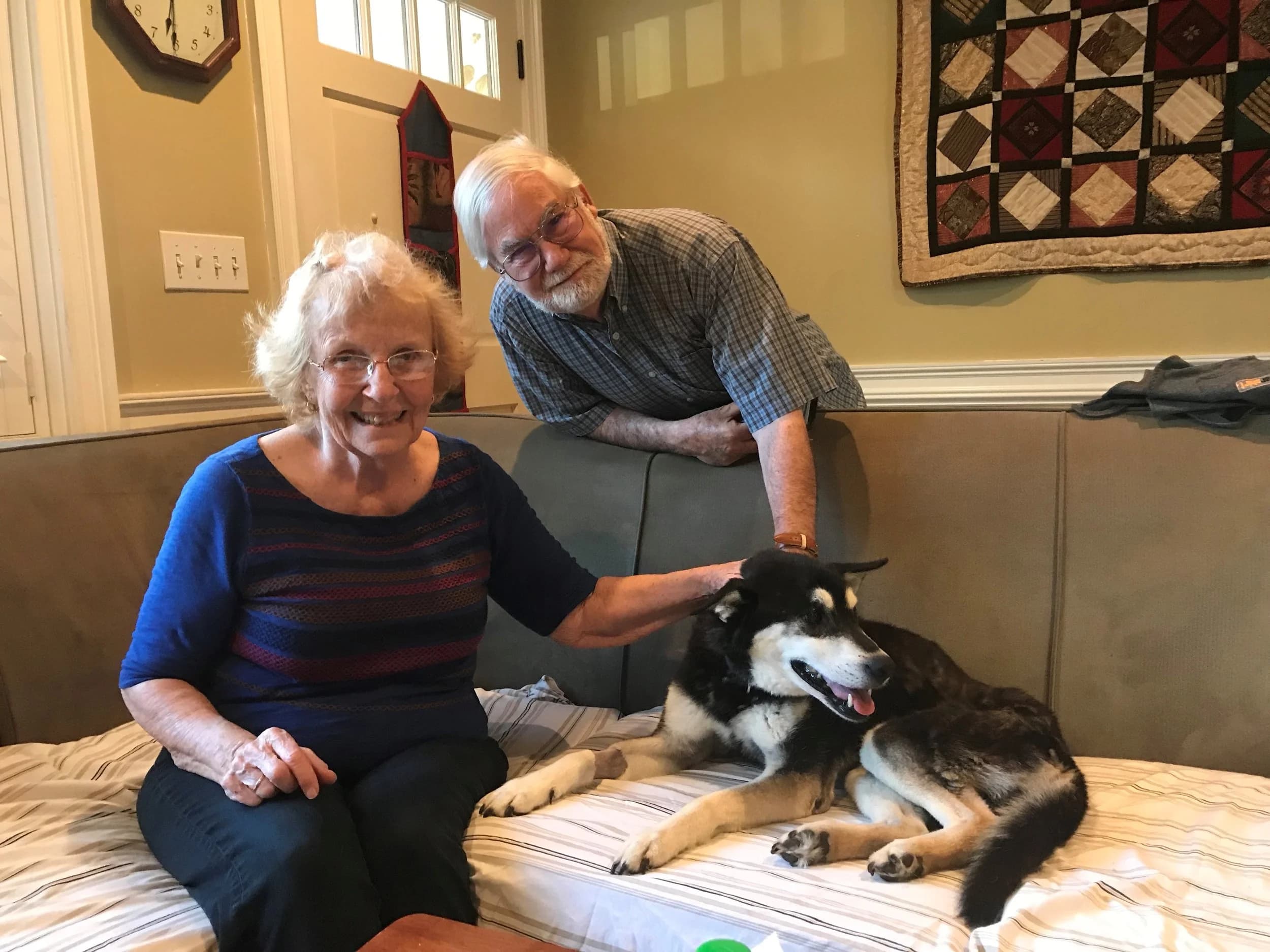 An elderly couple sits on a couch smiling warmly while petting a happy black and tan Husky