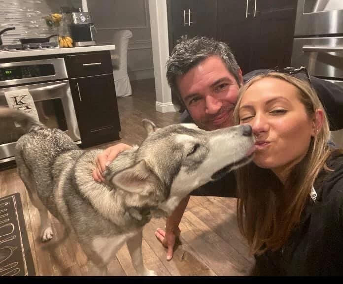 A couple smiling in a kitchen as a grey Husky gives one of them a kiss on the lips
