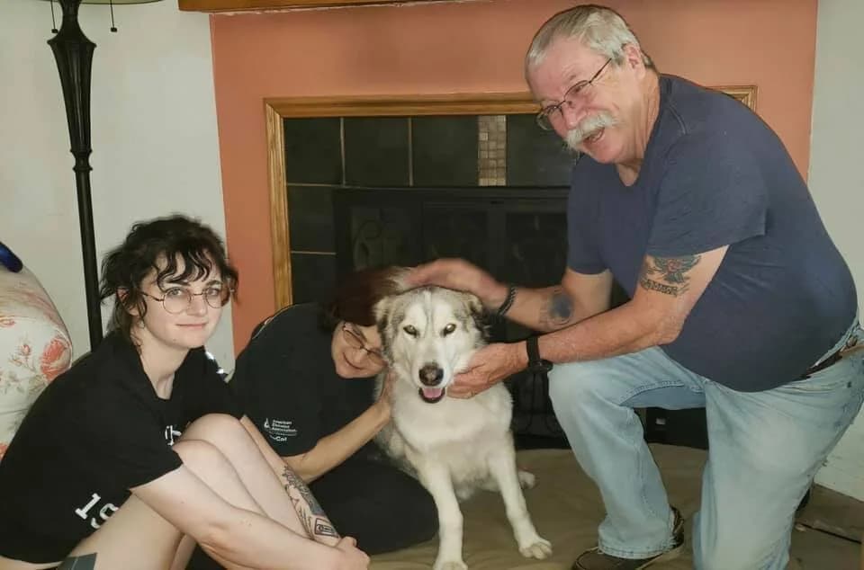 Two young adults and an older man smile while petting a husky dog indoors near a fireplace
